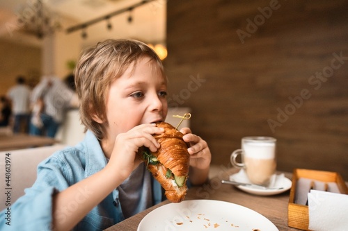 Happy cute boy eating fresh croissant  sitting at the table in city cafe  with paper cup of cocoa. High quality photo