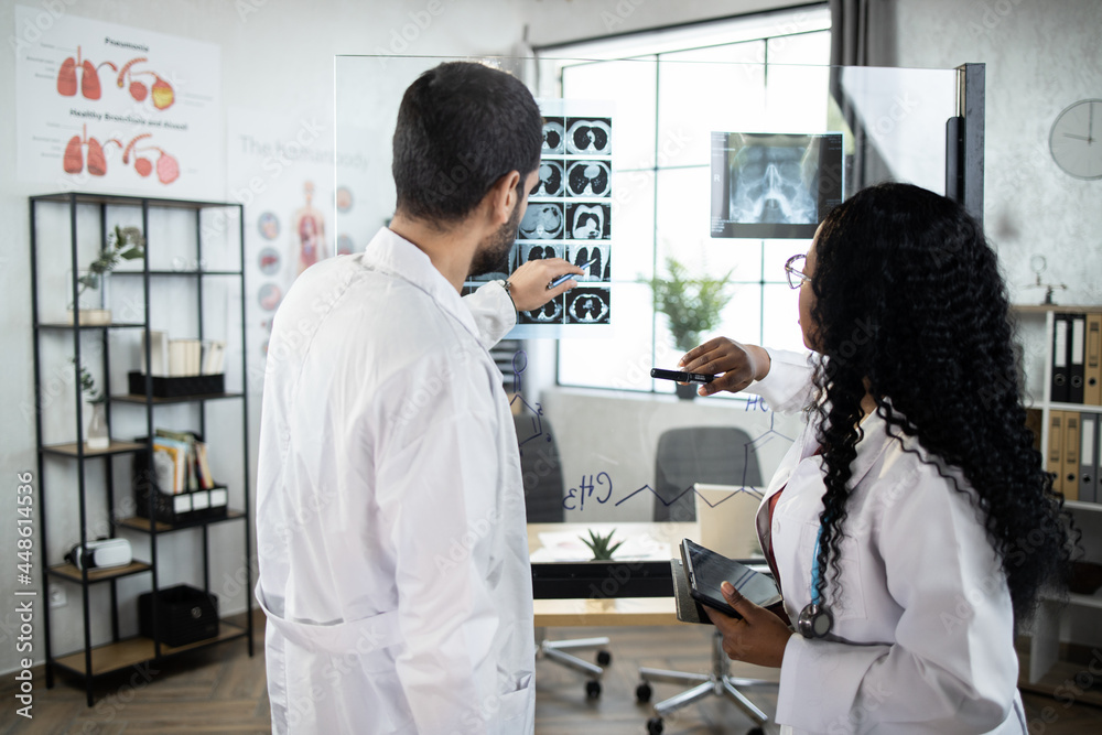 Two qualified multiethnic doctors, Arabian man and Afro-American woman, standing near the office glass board and interpreting x-ray image and CT scan of patient. Back view Two qualified multiethnic doctors, Arabian man and Afro-American woman, standing near the office glass board and interpreting x-ray image and CT scan of patient. Back view