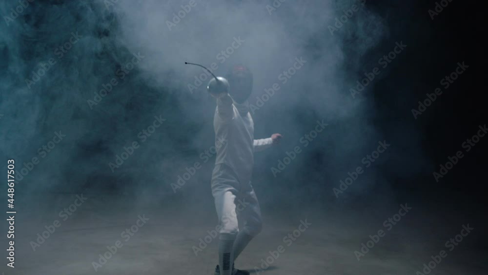 Professional fencer in protective uniform practicing maneuvers with sword alone in dark studio . Fencing demonstration on black smoked background . Shot on Arri Alexa cinema camera in slow motion . Professional fencer in protective uniform practicing maneuvers with sword alone in dark studio . Fencing demonstration on black smoked background . Shot on Arri Alexa cinema camera in slow motion .