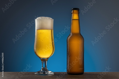 Unopened cold beer bottle and full beer glass on a wooden table blue background