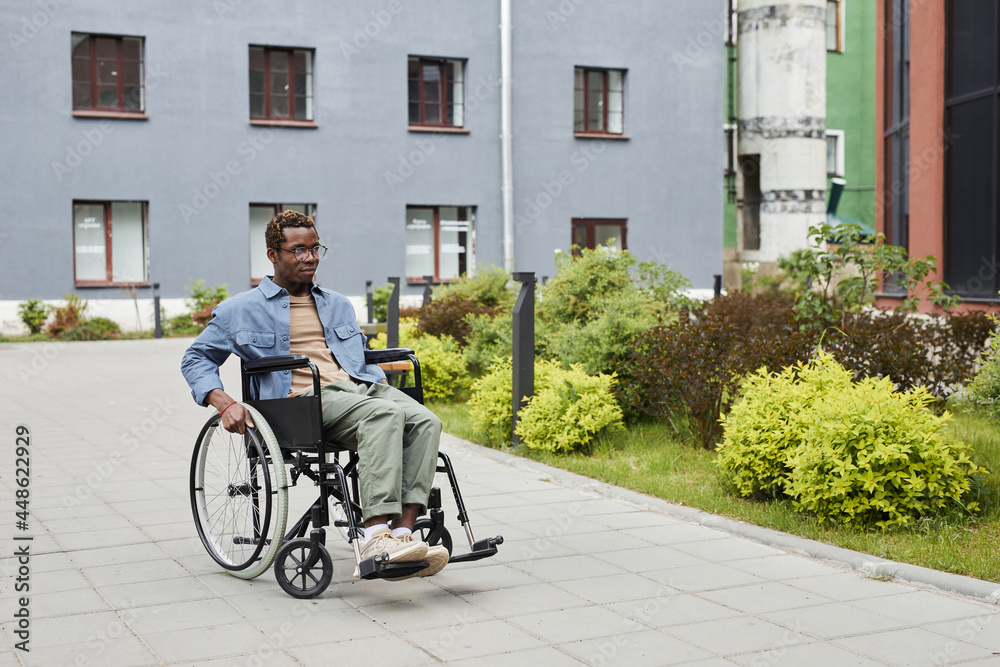 Serious handicapped young African-American man in eyeglasses sitting in wheelchair outdoors
