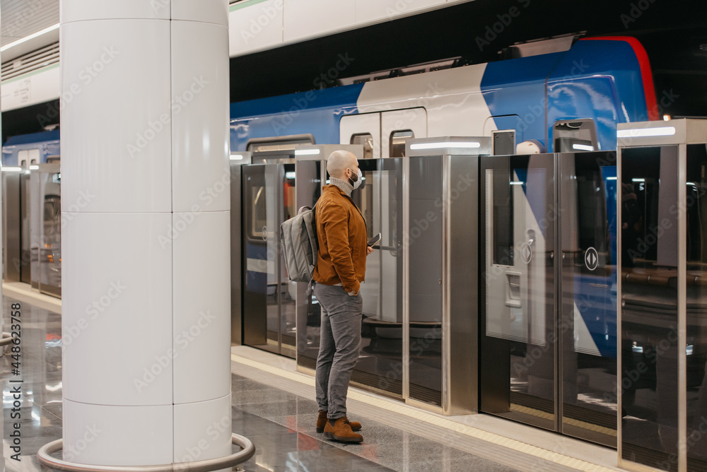 A man in a face mask is holding a smartphone while waiting for a subway train. A man in a face mask is holding a smartphone while waiting for a subway train.