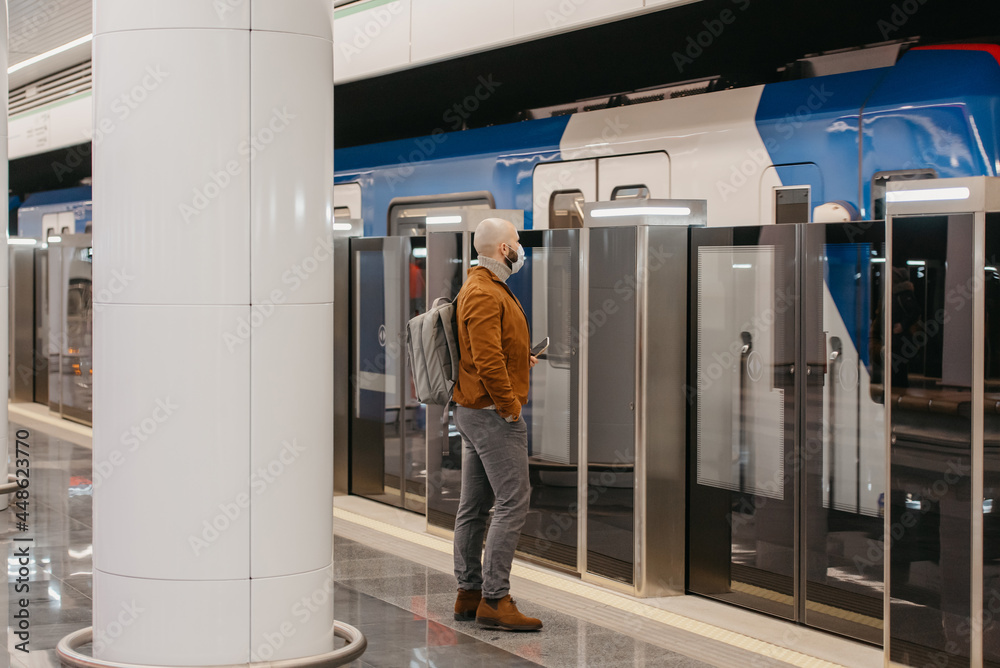 A man in a face mask is holding a smartphone while waiting for a subway train. A man in a face mask is holding a smartphone while waiting for a subway train.