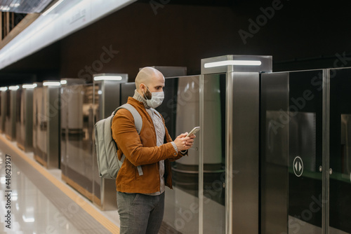 A man in a face mask is using a smartphone while waiting for a subway train.