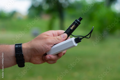 A man holds a power bank in his hands and charges an electronic cigarette against the backdrop of nature.