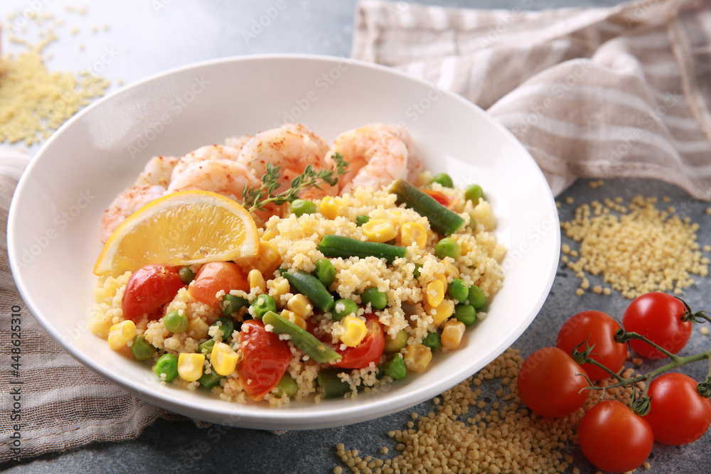 Seafood dish. Shrimp with couscous and vegetables: asparagus beans, corn, tomatoes, green peas and lemon in a white plate on grey table. Background image, copy space Seafood dish. Shrimp with couscous and vegetables: asparagus beans, corn, tomatoes, green peas and lemon in a white plate on grey table. Background image, copy space
