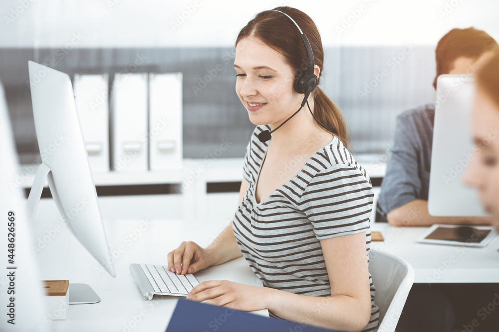 Obraz premium Casual dressed young woman using headset and computer while talking with customers online. Group of operators at work in sunny office. Call center