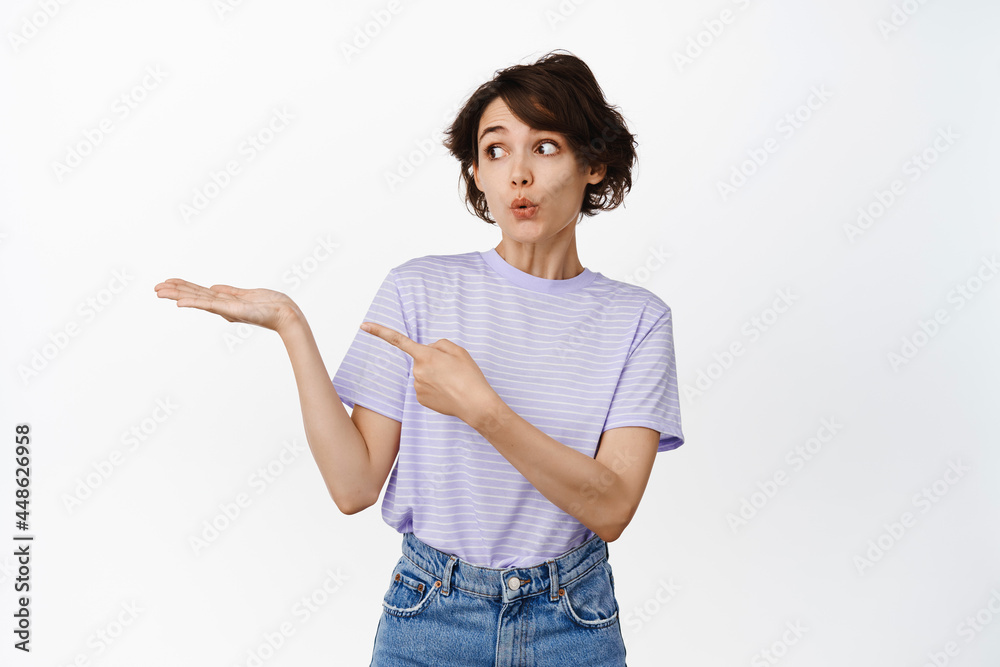 Surprised brunette woman pointing at her open hand, showing product, sale discount object on palm, standing in casual tshirt against white background Surprised brunette woman pointing at her open hand, showing product, sale discount object on palm, standing in casual tshirt against white background