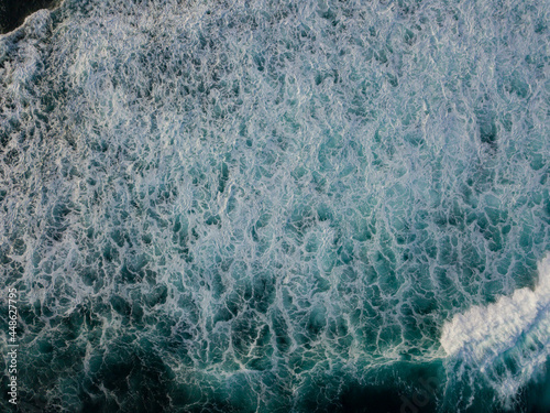 Drone photo of The dramatic lapping of the waves in the ocean produces white foam. The sea water of the pond is blue and contrasts with the color of the foam