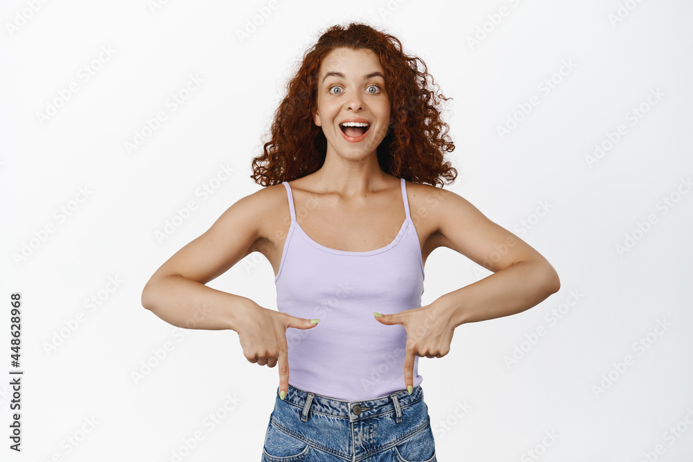 Excited curly-haired woman with red hair, pointing fingers down and smiling amazed, checking out, showing announcement, big news, standing over white background Excited curly-haired woman with red hair, pointing fingers down and smiling amazed, checking out, showing announcement, big news, standing over white background