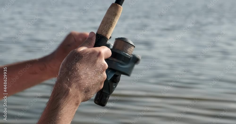 Spin fishing reel. Hands of a fisherman with a spinning rod in hand closeup. Handle rotation with reel of rod. Fisherman catches a fish, fishing on fishing rod spinning. Calm surface water. Close up