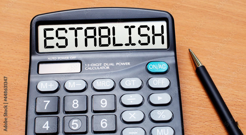 On a wooden table there is a black pen and a calculator with the text ESTABLISH. Business concept On a wooden table there is a black pen and a calculator with the text ESTABLISH. Business concept