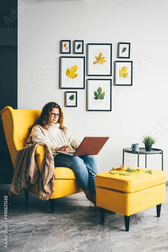 Young woman with long loose curly hair sits in armchair and types on coloured laptop looking into display and smiling against designed wall at home.