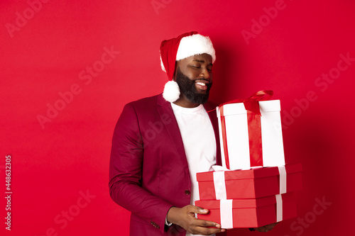 Christmas, New Year and shopping concept. Happy african amaerican man looking touched and thankful at xmas presents, receiving gifts, wearing santa hat, red background photo