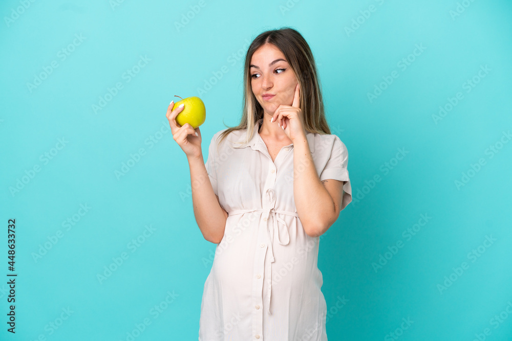 Young Romanian woman isolated on blue background pregnant and holding an apple while thinking