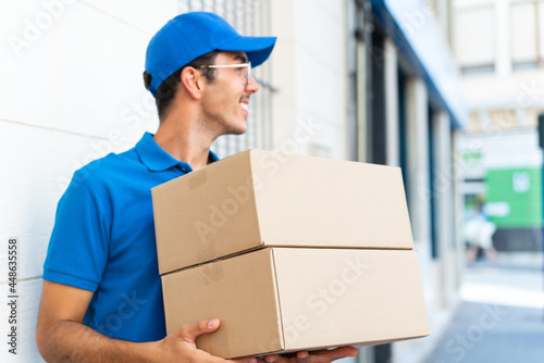 Young delivery man at outdoors holding boxes with happy expression looking side