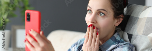 Fotografija Surprised woman looks at smartphone screen while sitting on couch