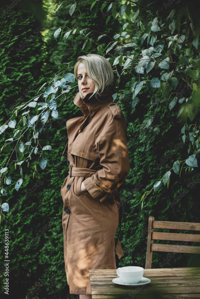 Beautiful woman in coat stay next to table with cup of coffee in a garden with trees on background Beautiful woman in coat stay next to table with cup of coffee in a garden with trees on background