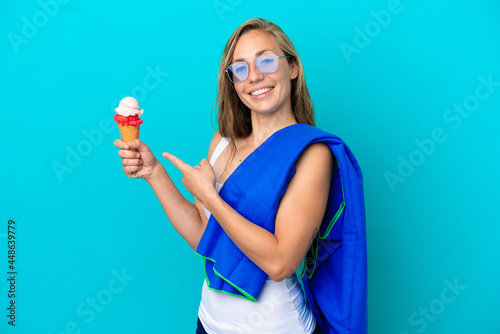 Young caucasian woman holding ice cream and wearing a beach towel isolated on blue background and pointing it