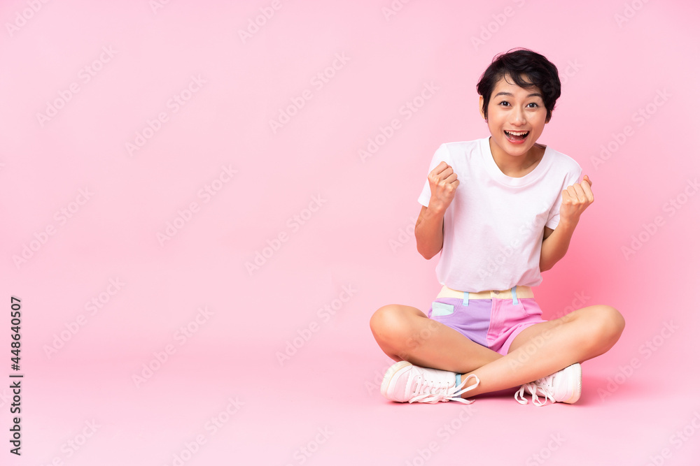 Young Vietnamese woman with short hair sitting on the floor over isolated pink background celebrating a victory in winner position Young Vietnamese woman with short hair sitting on the floor over isolated pink background celebrating a victory in winner position