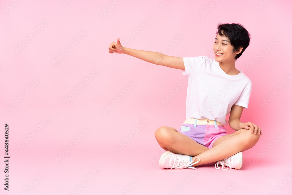 Young Vietnamese woman with short hair sitting on the floor over isolated pink background giving a thumbs up gesture Young Vietnamese woman with short hair sitting on the floor over isolated pink background giving a thumbs up gesture