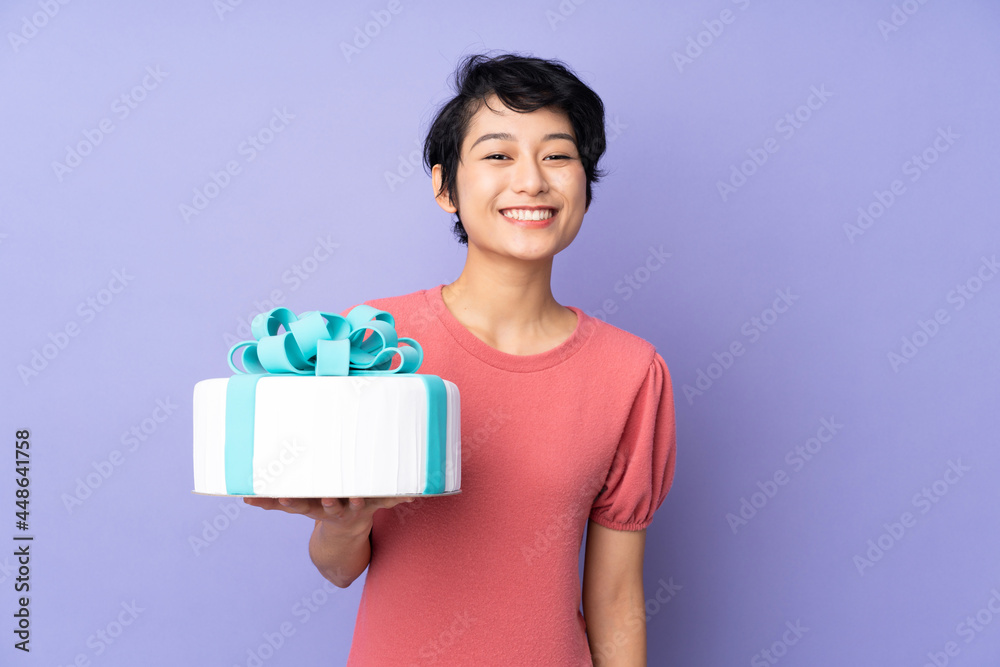 Young Vietnamese woman with short hair holding a big cake over isolated purple background smiling a lot Young Vietnamese woman with short hair holding a big cake over isolated purple background smiling a lot