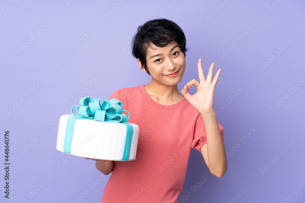 Young Vietnamese woman with short hair holding a big cake over isolated purple background showing ok sign with fingers Young Vietnamese woman with short hair holding a big cake over isolated purple background showing ok sign with fingers