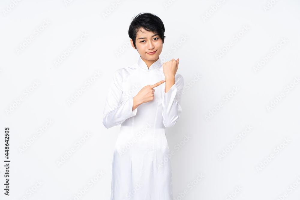 Young Vietnamese woman with short hair wearing a traditional dress over isolated white background making the gesture of being late Young Vietnamese woman with short hair wearing a traditional dress over isolated white background making the gesture of being late