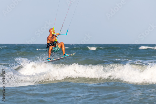 Young adult caucasian fit male person enjoy riding kite surf board making extreme trick stunt bright sunny day against blue sky at sea or ocean shore. Watersport adrenaline fun adventure acitivity