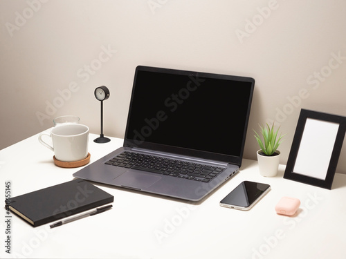 Picture of laptop on a white table with a mug a clock a small bush a notebook a phone a picture frame and a headphone case