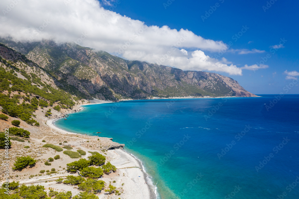 Aerial view of clouds over large mountains on a coastline surrounded by clear, blue ocean (Agia Roumeli, Crete)
