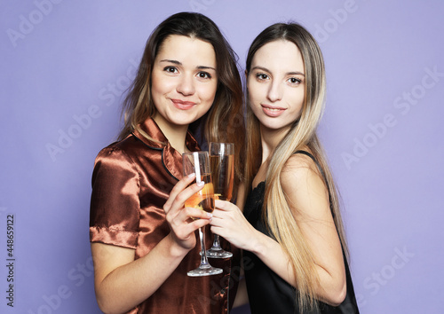 two beautiful women dressed in pajamas drink champagne and have fun over purple background
