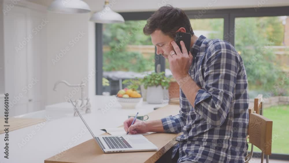 Man working from home using laptop on kitchen counter talking on mobile phone- shot in slow motion