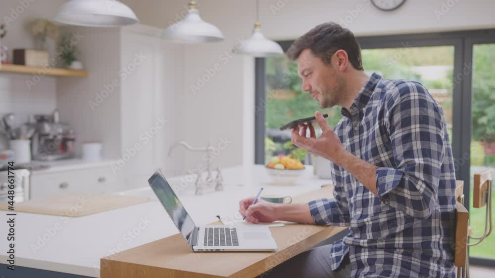 Man working from home using laptop on kitchen counter talking into microphone of mobile phone- shot in slow motion Man working from home using laptop on kitchen counter talking into microphone of mobile phone- shot in slow motion