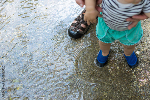 Father holding toddler daughter's hands as they walk through a shallow stream; water shoes