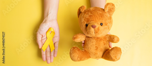 hand holding Yellow Ribbon and Bear doll on yellow background for supporting kid living and illness. September Childhood Cancer Awareness month and World cancer day concept