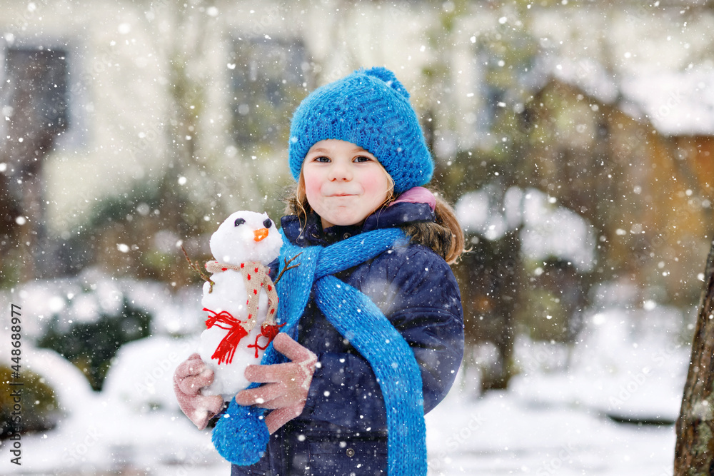 Cute little toddler girl making mini snowman and eating carrot nose. Adorable healthy happy child playing and having fun with snow, outdoors on cold day. Active leisure with children in winter Cute little toddler girl making mini snowman and eating carrot nose. Adorable healthy happy child playing and having fun with snow, outdoors on cold day. Active leisure with children in winter