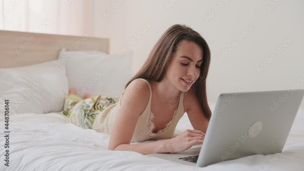 Smiling fun young brunette woman in pajamas beige tank-top lying in bed hold use work on laptop pc computer spend time in bedroom lounge home in own room house. Rest relax good mood lifestyle concept Smiling fun young brunette woman in pajamas beige tank-top lying in bed hold use work on laptop pc computer spend time in bedroom lounge home in own room house. Rest relax good mood lifestyle concept