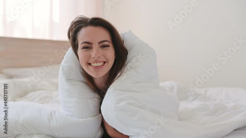 Happy brunette young woman in casual beige tank-top pajamas lying in bed wrapped in blanket look camera relax spend time in bedroom lounge home in own room house. Bedtime good mood lifestyle concept