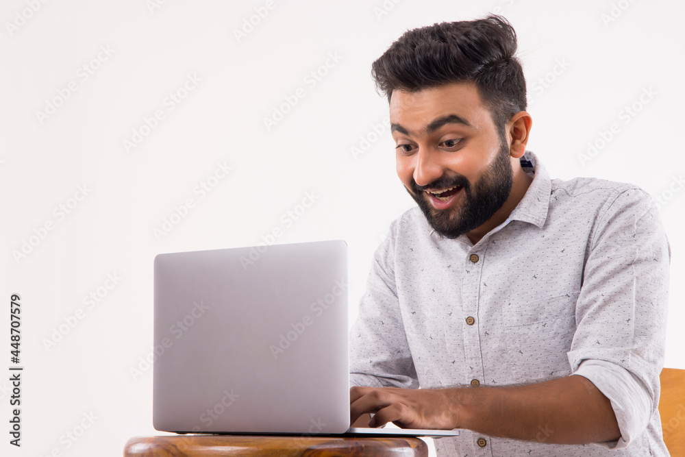 A BEARDED YOUNGSTER LOOKING AT LAPTOP IN EXCITEMENT A BEARDED YOUNGSTER LOOKING AT LAPTOP IN EXCITEMENT