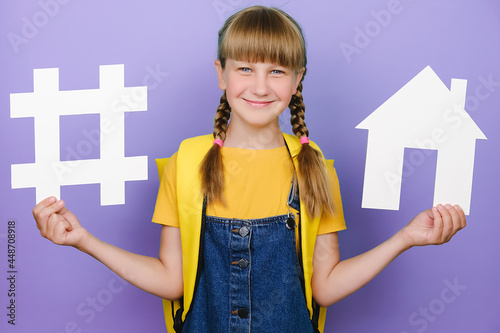 Happy blonde schoolgirl holding white hashtag sign and house model  label for business  wears yellow backpack  posing over purple background. Social network monitoring  media measurement  school