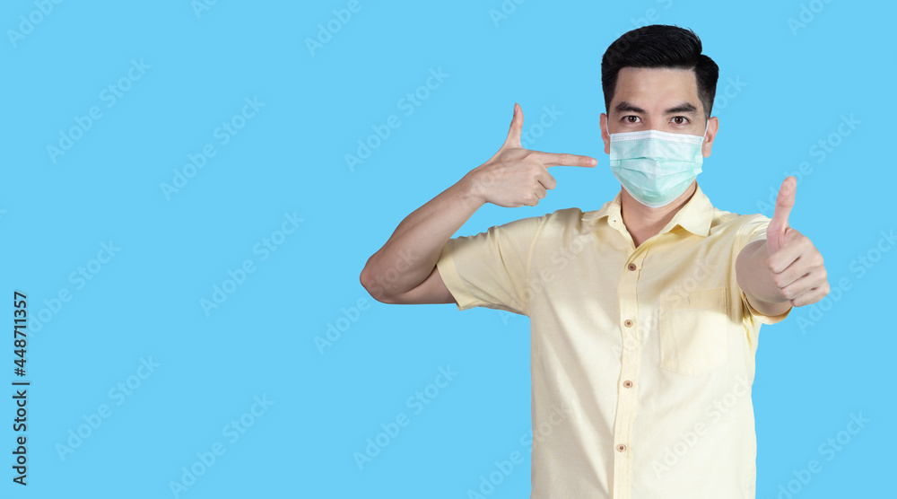 COVID-19 Coronavirus portrait handsome young asian man wearing yellow shirt and mask protection from covid 19 and thumbs up sign isolated on blue background in studio. Asian people. COVID-19 concept. COVID-19 Coronavirus portrait handsome young asian man wearing yellow shirt and mask protection from covid 19 and thumbs up sign isolated on blue background in studio. Asian people. COVID-19 concept.