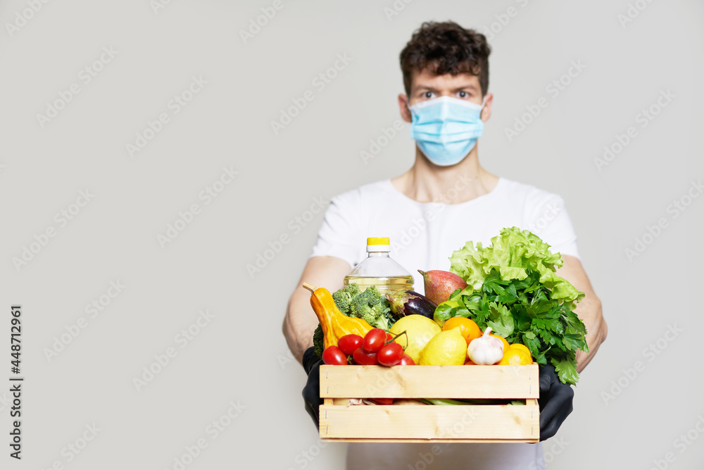 Food delivery concept during quarantine. A man in a mask, gloves and a white T-shirt holds a box of fresh food in his hands. Food delivery from the supermarket, from farmers home. Food delivery concept during quarantine. A man in a mask, gloves and a white T-shirt holds a box of fresh food in his hands. Food delivery from the supermarket, from farmers home.