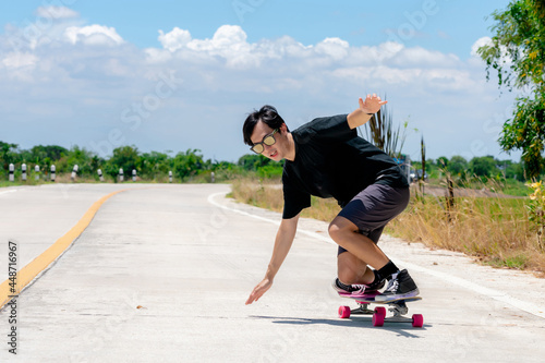 A young Asian man is wearing a black shirt and pants. play skateboard Show the posture of a turn around. On a country road on a sunny day with sky. Looking at the camera, Play surf skate. photo