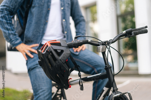 Close-up cropped shot of bicycle helmet hanging on handlebars of bike standing in city street summer day, on blurry background of urban building. Unrecognizable cyclist sitting on seat of bike.