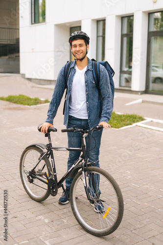 Fototapeta Naklejka Na Ścianę i Meble -  Vertical portrait of cheerful handsome young delivery man in protective helmet posing standing near bicycle in city street, looking at camera. Courier male with thermo backpack delivery food to client