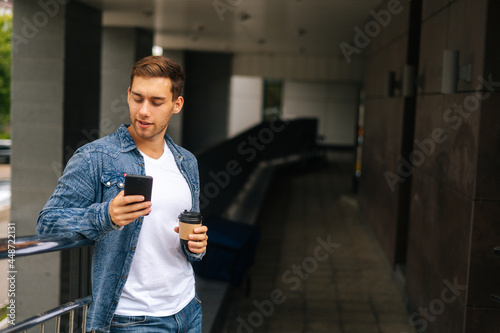 Medium shot portrait of confident young man using typing mobile phone, standing on balcony terrace with coffee coffee cup leaning on railing of office building, blurred background.