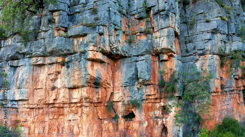 Red rugged cliff - sedimentary rock mountain formation. Vertical natural rough wall of stratum layers, colorful, sliced and cracked, brown, orange color due to ferric oxides. Geological strata canyon.