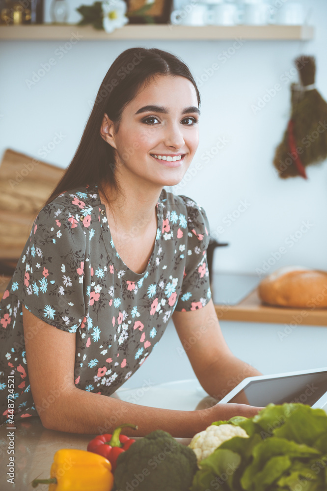 An attractive young dark-haired woman chooses the recipe for a delicious meal while sitting at the table in the kitchen. Tablet pc is the best cookbook An attractive young dark-haired woman chooses the recipe for a delicious meal while sitting at the table in the kitchen. Tablet pc is the best cookbook