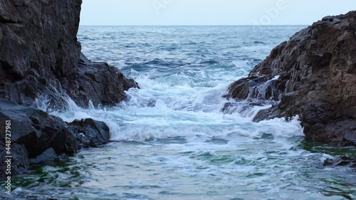 Gentle blue sea waves rushing and splashing between dark cliffs forming white foam. Rocks on both sides